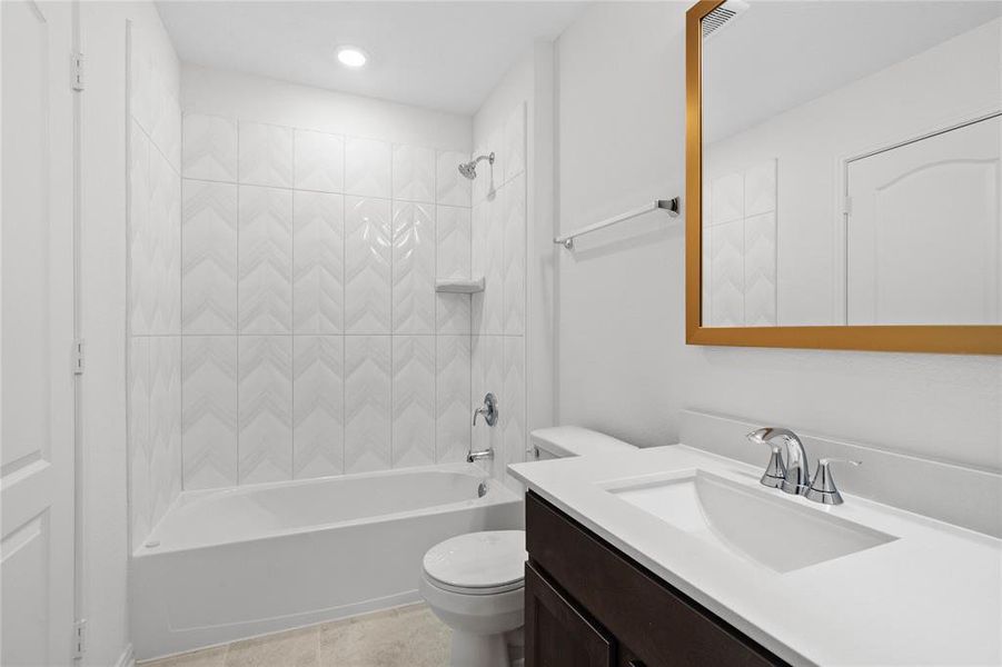 Secondary bath features tile flooring, bath/shower combo with tile surround, stained wood cabinets, beautiful light countertops, mirror, dark, sleek fixtures and modern finishes.