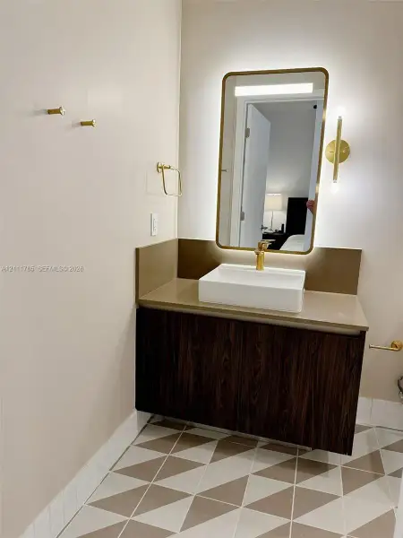 Floating vanity with wood-finish cabinetry, rectangular vessel sink, brass-tone hardware, backlit wall mirror, and geometric patterned floor tiling