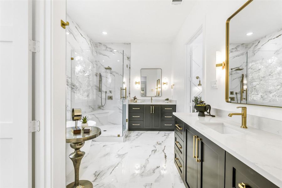 A breathtaking overview of the primary ensuite reveals a harmonious blend of luxury materials and thoughtful design. The space features 24x48polished porcelain marble tile extending from floor to ceiling, complemented by custom dual vanities finished in rich charcoal cabinetry with soft-close drawers and brushed brass pulls. Gold-framed mirrors and matching sconces add a warm glow, while the frameless glass shower enclosure showcases floor-to-ceiling tile and designerfixtures.