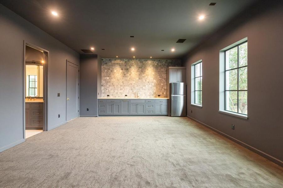 This stylish media room boasts dark walls, plush carpeting, and a built-in kitchenette with sink, full refrigerator, contemporary cabinetry, and a sleek backsplash. It's equipped with ample lighting and large windows for a cozy yet bright entertainment space.