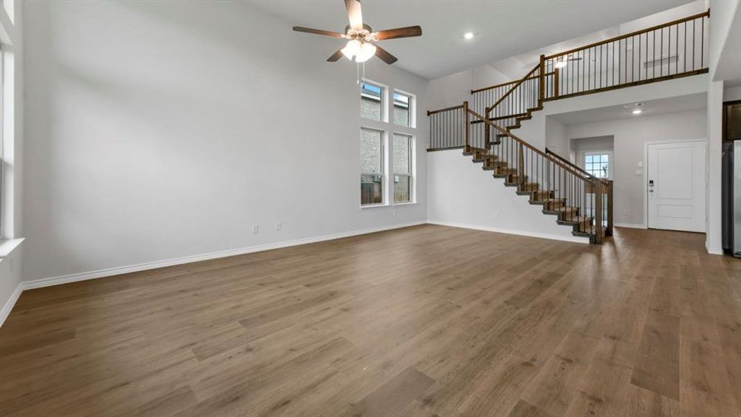 Spacious living area featuring high ceilings, wood-finish flooring, a contemporary ceiling fan, and a prominent staircase with dark wood handrails and black iron balusters