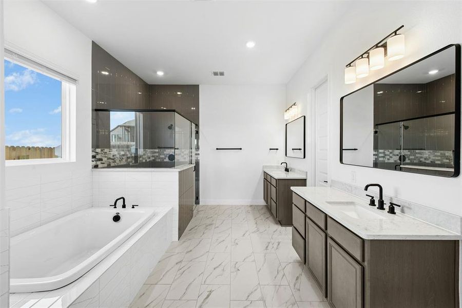 Spacious bathroom featuring a built-in soaking tub with white tile surround, a separate glass-enclosed shower with dark tile walls, dual vanities with light countertops and dark wood-finish cabinetry, and large rectangular framed mirrors with contemporary lighting fixtures
