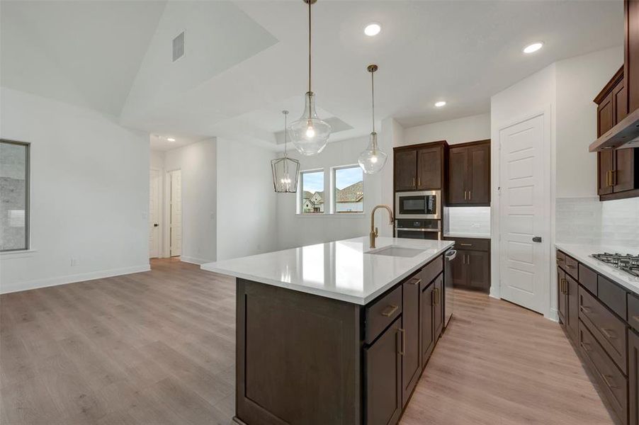Open-concept kitchen featuring a central island with a white countertop and integrated sink, pendant lighting, dark wood cabinetry with gold hardware, stainless steel appliances, and wood-finish flooring