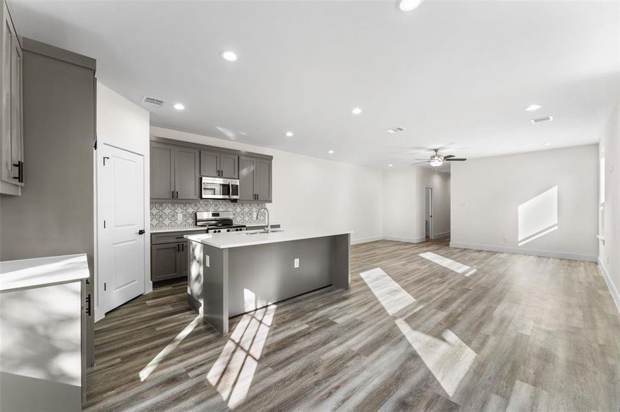 Kitchen featuring a kitchen island with sink, appliances with stainless steel finishes, gray cabinetry, ceiling fan, and dark wood-style flooring Kitchen featuring a kitchen island with sink, appliances with stainless steel finishes, gray cabinetry, ceiling fan, and dark wood-style flooring