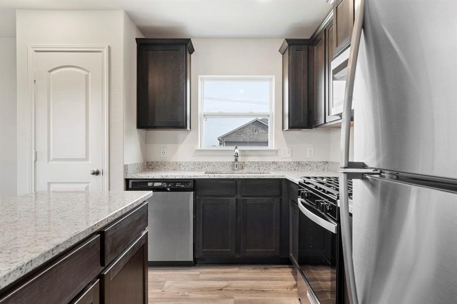 Kitchen featuring light stone countertops, light wood-type flooring, dark brown cabinetry, sink, and appliances with stainless steel finishes