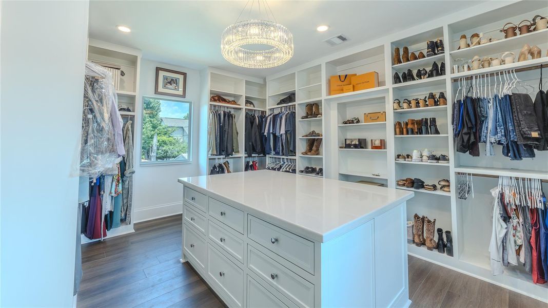 This spacious walk-in closet features custom shelving, a large central island with drawers, and ample hanging space. A window provides natural light, and a modern chandelier adds elegance to the room. Perfect for organized storage and a stylish dressing experience.