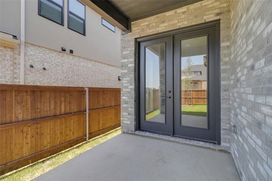Concrete patio with grey brick exterior, featuring double glass-paneled doors, wood privacy fencing, and green lawn space