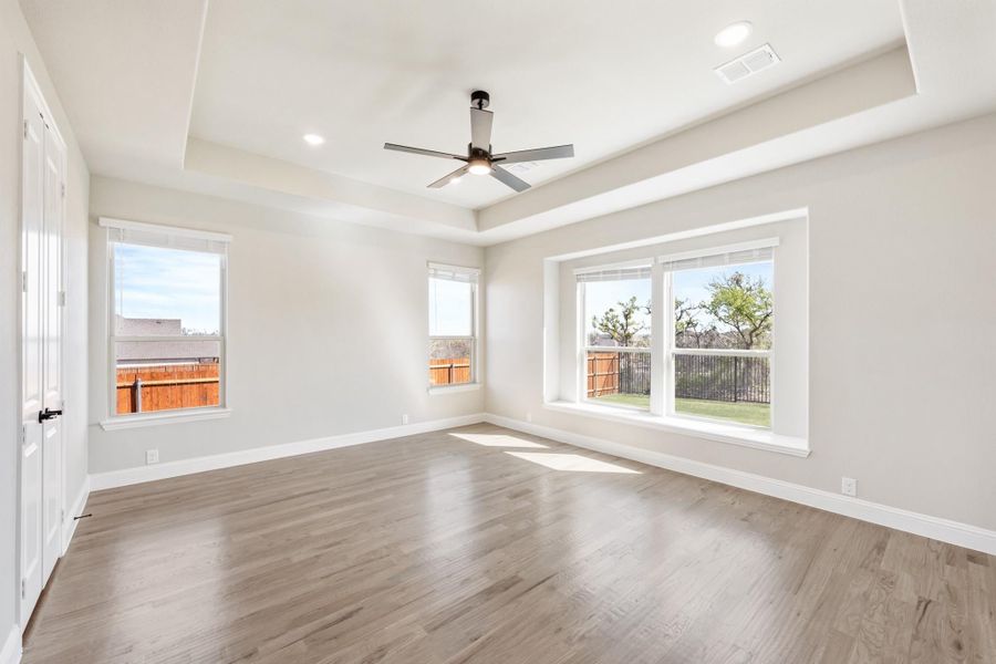 Empty bedroom with tray ceiling, ceiling fan, hardwood floors, and three large windows with backyard views