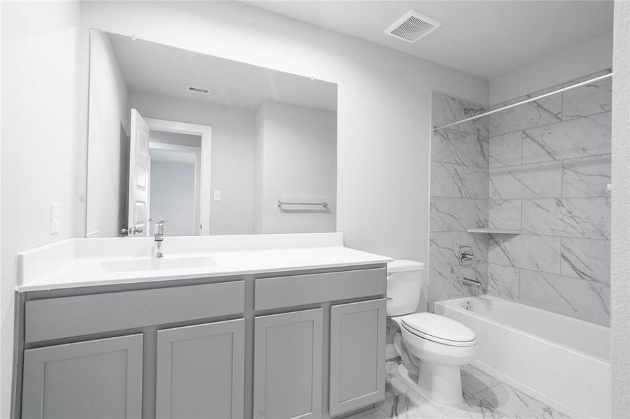 Secondary bath features tile flooring, bath/shower combo with tile surround, stained wood cabinets, beautiful light countertops, mirror, dark, sleek fixtures and modern finishes.