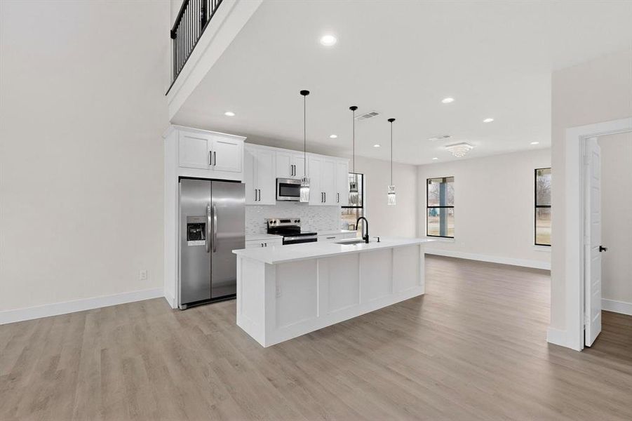Kitchen with stainless steel appliances, white cabinetry, a center island with sink, open floor plan, and decorative light fixtures Kitchen with stainless steel appliances, white cabinetry, a center island with sink, open floor plan, and decorative light fixtures