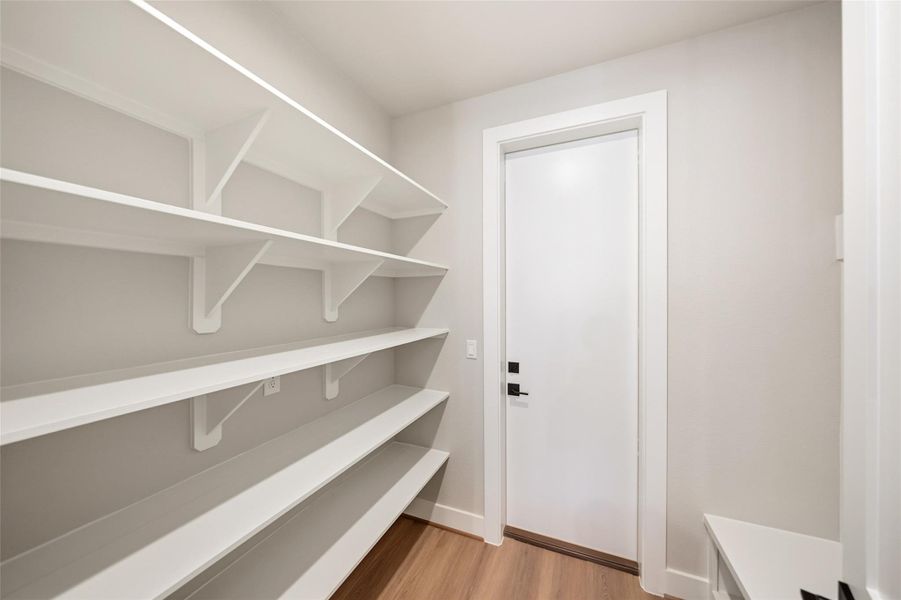 Spacious pantry with white shelving on the left and a sleek white door straight ahead. The room features light walls and wood flooring, ideal for organized storage.