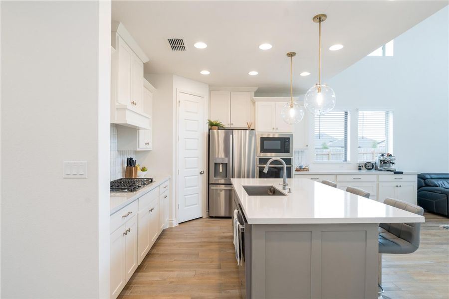 Dual tone kitchen featuring stainless steel appliances, a kitchen island with sink, a kitchen bar, light wood-style flooring, and decorative light fixtures