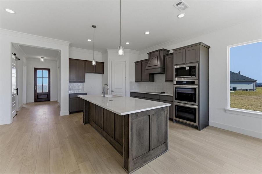 Kitchen with a kitchen island with sink, stainless steel appliances, decorative light fixtures, backsplash, and light wood-style flooring