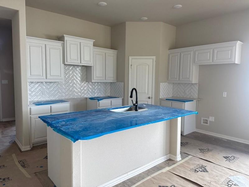 Kitchen island with an undermount sink and a matte black faucet, surrounded by white cabinetry, a herringbone pattern tile backsplash, and recessed ceiling lighting Kitchen island with an undermount sink and a matte black faucet, surrounded by white cabinetry, a herringbone pattern tile backsplash, and recessed ceiling lighting