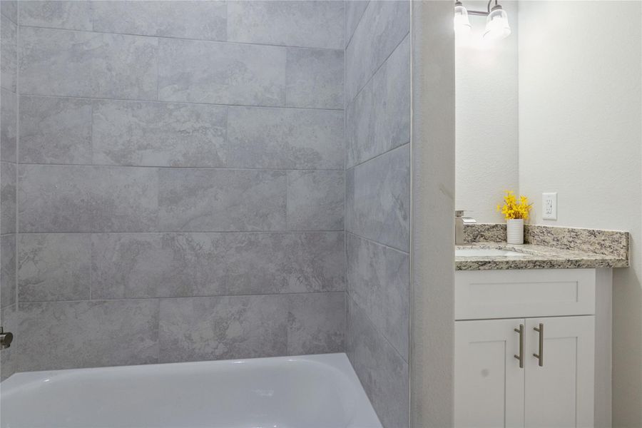 This bathroom features a modern design with gray tiled walls and a white bathtub. The vanity has a granite countertop, white cabinetry, and is accented by bright lighting and a small vase of yellow flowers.
