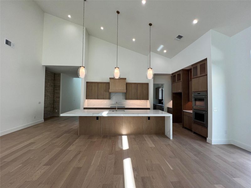 Kitchen featuring a large island with sink, light stone counters, decorative light fixtures, stainless steel double oven, and light wood-style floors