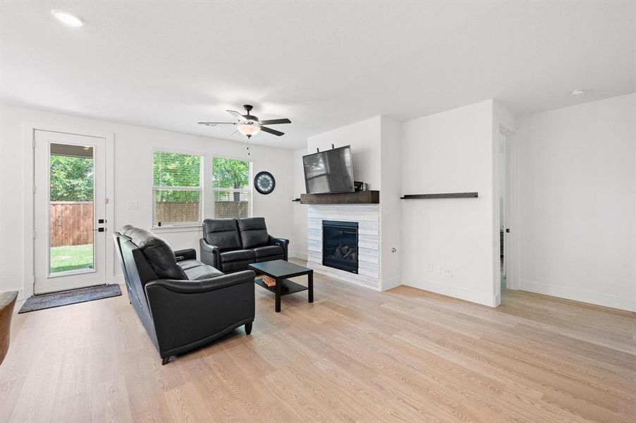 Spacious living area featuring light wood-finish flooring, a contemporary fireplace with a white tile surround and wood mantel, and a ceiling fan Spacious living area featuring light wood-finish flooring, a contemporary fireplace with a white tile surround and wood mantel, and a ceiling fan