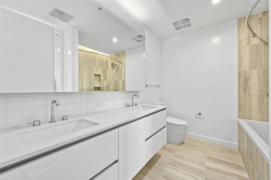 The primary bathroom features a sleek, modern design with a dual vanity, offering ample space for two. The clean white cabinetry, paired with matching countertops, enhances the room's contemporary aesthetic. The large mirror above the vanity adds a sense of openness, while the integrated lighting provides a bright and inviting atmosphere. The flooring complements the space with a warm wood-like texture, and the shower & tub, visible in the background, features stylish tiling for a luxurious feel.