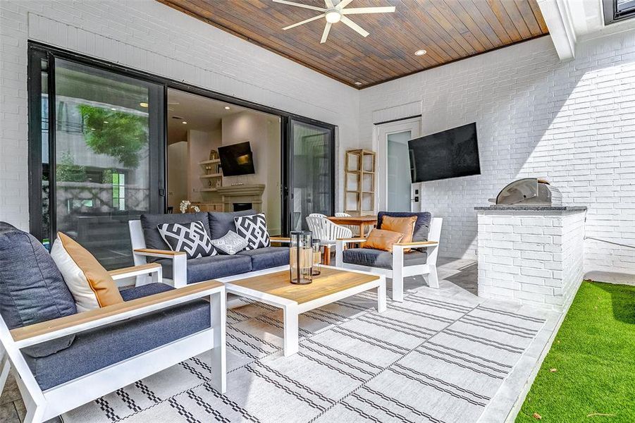 This inviting outdoor patio features a cozy seating area with modern furniture, a ceiling fan, and a mounted TV. The space is complemented by a built-in grill and white brick walls, offering a perfect setting for relaxation and entertaining.