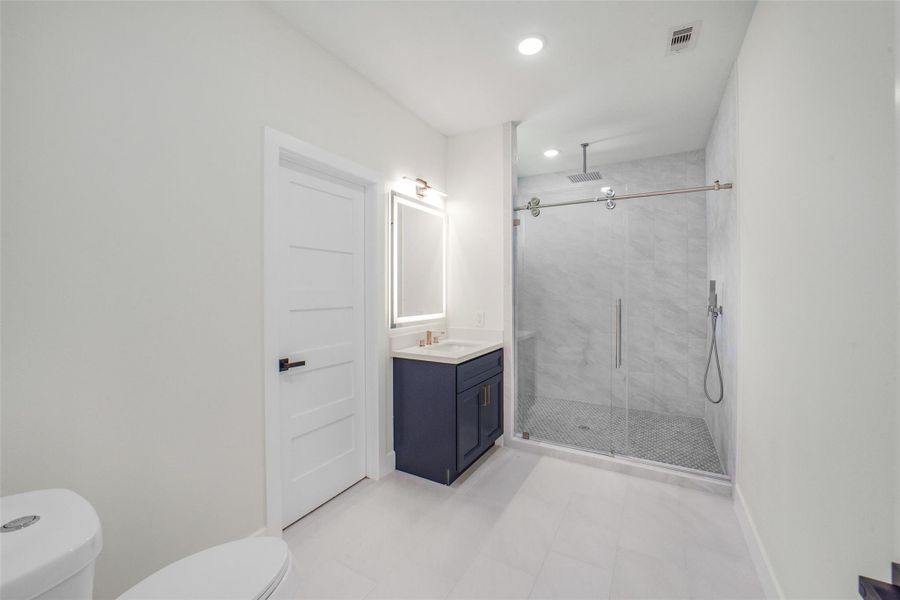This photo shows a modern bathroom featuring a spacious glass-enclosed shower with sleek fixtures, a dark vanity with a lighted mirror, and bright white walls and flooring for a clean, contemporary look.