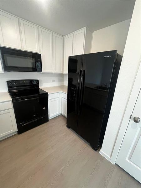 Kitchen featuring white shaker cabinetry, light-toned countertops, contemporary black appliances, and wood-finish flooring Kitchen featuring white shaker cabinetry, light-toned countertops, contemporary black appliances, and wood-finish flooring