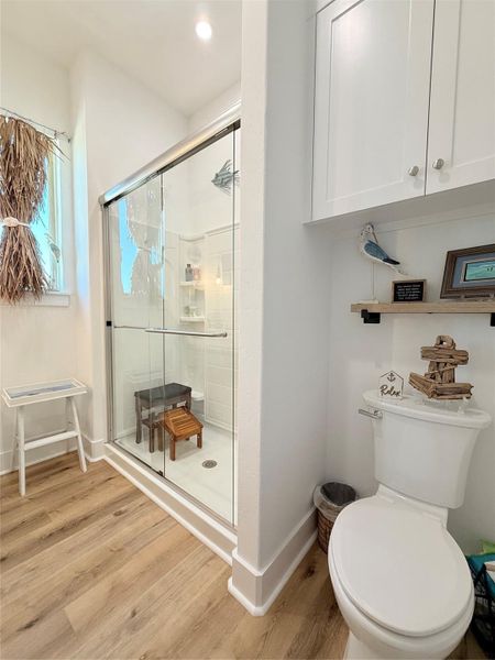 This is a bright, modern bathroom featuring a glass-enclosed shower, white walls, and light wood flooring. It has a cozy, decorative touch with shelving above the toilet and a small window for natural light.