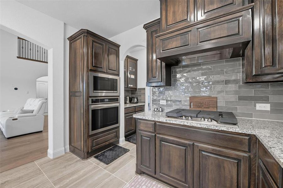 Kitchen featuring dark brown cabinetry, decorative backsplash, stainless steel appliances, arched walkways, and light stone counters Kitchen featuring dark brown cabinetry, decorative backsplash, stainless steel appliances, arched walkways, and light stone counters