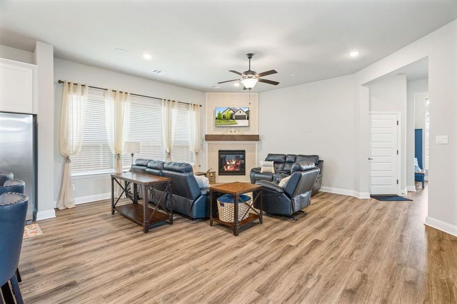 Spacious living area featuring wood-finish flooring, a corner fireplace with a stone surround and wood mantel, multiple windows with blinds, a ceiling fan with integrated lighting, and recessed ceiling lighting Spacious living area featuring wood-finish flooring, a corner fireplace with a stone surround and wood mantel, multiple windows with blinds, a ceiling fan with integrated lighting, and recessed ceiling lighting
