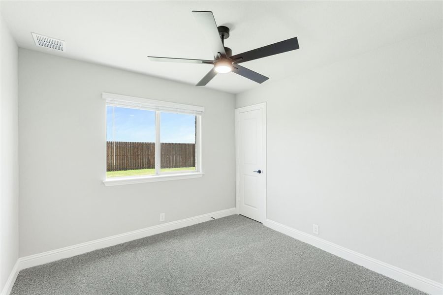 Empty bedroom with gray carpet, ceiling fan, and single window overlooking backyard fence
