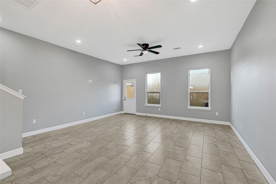 This spacious, open-concept living area features neutral gray walls, tile flooring, recessed lighting, and a ceiling fan. It has two large windows and a door leading outside, offering a bright and inviting atmosphere.