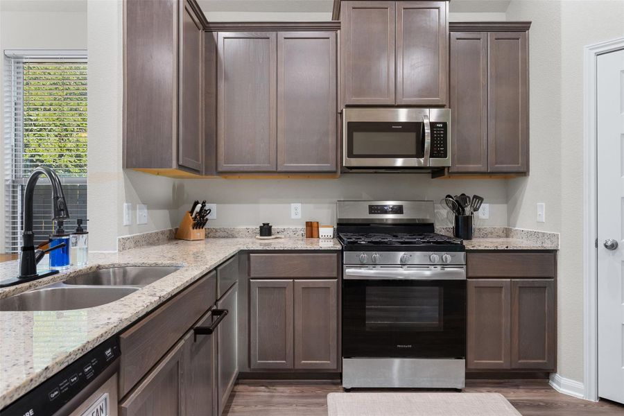 This modern kitchen features sleek dark wood cabinets, granite countertops, and stainless steel appliances, including a gas stove and microwave. Also includes a double sink with a stylish faucet.