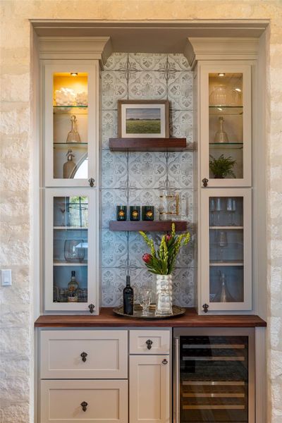 Indoor dry bar with wooden counters, glass insert cabinets, wine cooler, and white cabinets