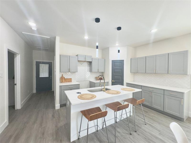 Kitchen featuring decorative light fixtures, light hardwood / wood-style floors, backsplash, a kitchen island with sink, and sink