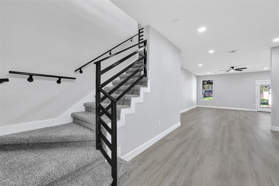 This photo showcases a modern, open-concept living area with light wood flooring and a sleek staircase featuring black metal railings. The space is bright, with recessed lighting and a ceiling fan, and includes a large window and a glass door leading outside.
