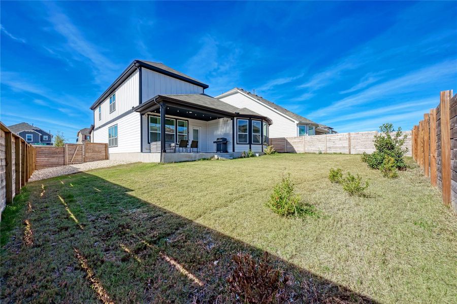 Expansive fenced backyard featuring a covered patio, a two-story home exterior with white siding and dark trim, a manicured lawn, and a gravel side yard with a wooden gate