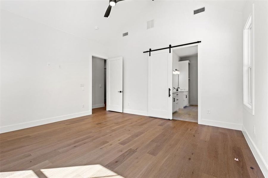 Unfurnished bedroom with a barn door, light wood-style floors, ceiling fan, ensuite bathroom, and high vaulted ceiling Unfurnished bedroom with a barn door, light wood-style floors, ceiling fan, ensuite bathroom, and high vaulted ceiling