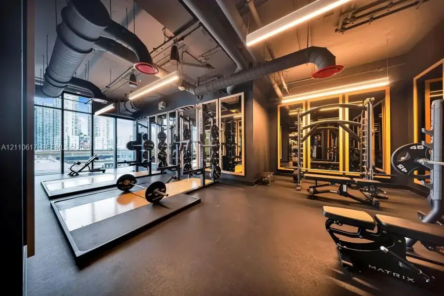 Fitness facility featuring rubber flooring, floor-to-ceiling windows, industrial-style exposed ductwork, wall-mounted mirrors, and integrated linear lighting Fitness facility featuring rubber flooring, floor-to-ceiling windows, industrial-style exposed ductwork, wall-mounted mirrors, and integrated linear lighting