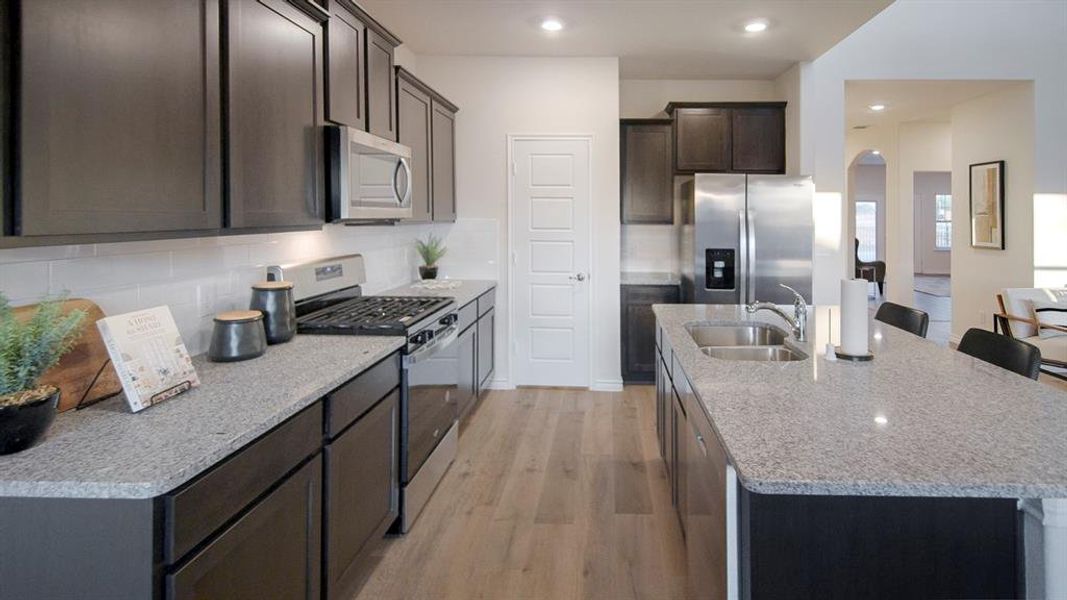 Modern kitchen featuring dark wood-finish cabinetry, light speckled countertops, stainless steel appliances, a tile backsplash, and light wood-finish flooring Modern kitchen featuring dark wood-finish cabinetry, light speckled countertops, stainless steel appliances, a tile backsplash, and light wood-finish flooring