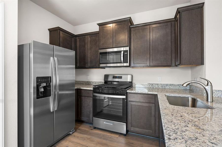 Kitchen with appliances with stainless steel finishes, light stone counters, dark brown cabinets, and dark wood-style flooring