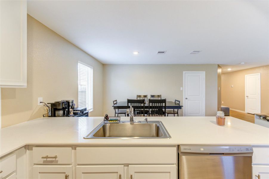 This photo showcases a bright, open kitchen with a modern design featuring a central island with a sink and dishwasher. It leads into a dining area with ample natural light, neutral walls, and recessed lighting, creating a welcoming space.