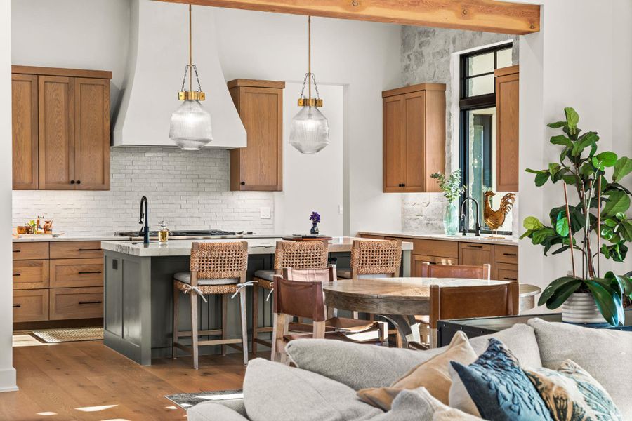 Kitchen with tasteful backsplash, custom range hood, brown cabinets, decorative light fixtures, and light stone counters Kitchen with tasteful backsplash, custom range hood, brown cabinets, decorative light fixtures, and light stone counters