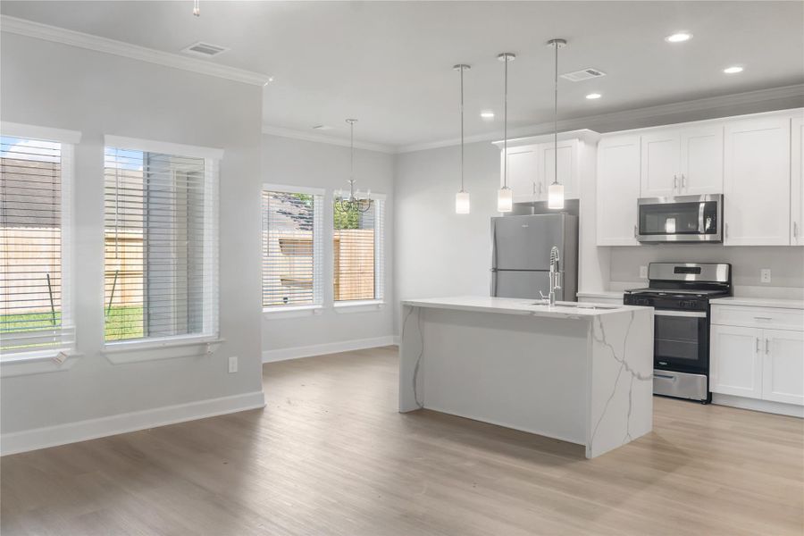 This photo showcases a bright, modern kitchen with white cabinetry, stainless steel appliances, and an island with pendant lighting. The open layout connects to a dining area with large windows, offering plenty of natural light and a view of the fenced backyard.