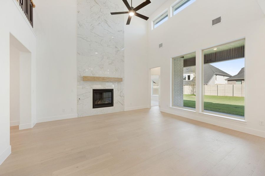 Open living room with two-story marble fireplace, wood floors, and large windows overlooking backyard