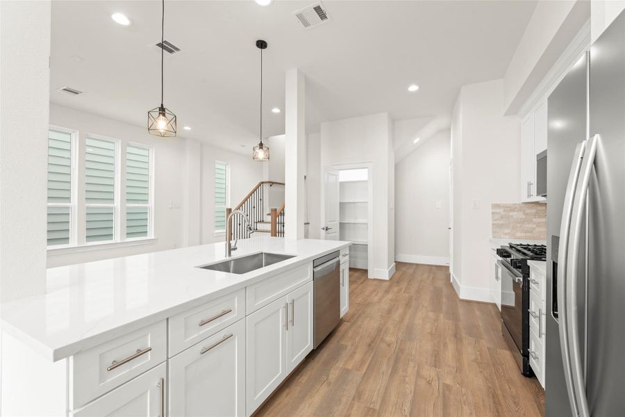 This kitchen features a modern design with sleek white cabinetry and a large island with a built-in sink. Stainless steel appliances, including a fridge and stove, complement the space. Pendant lighting adds elegance. An open layout leads to a staircase and a pantry area.