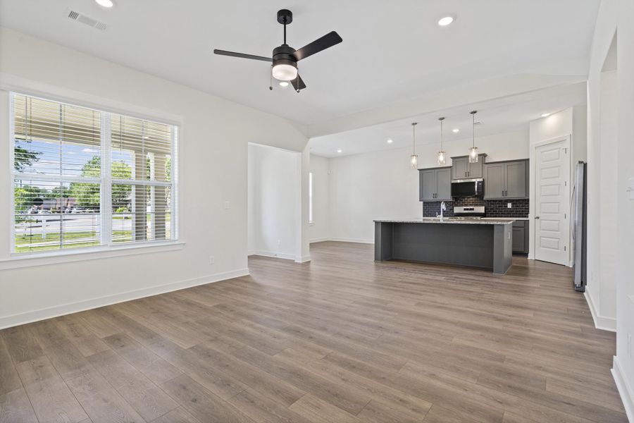Open-concept living space with wood-finish flooring, a large window with horizontal blinds, and a ceiling fan with integrated lighting