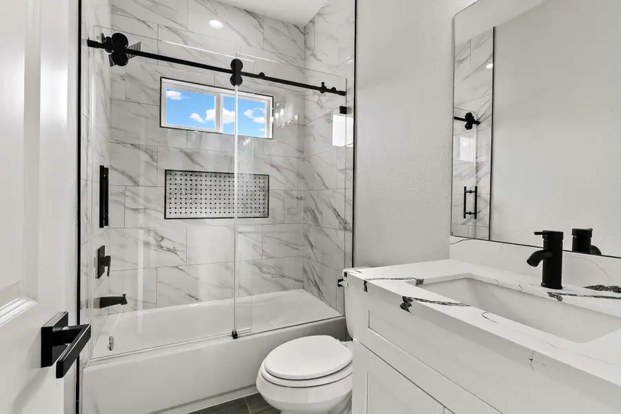 This bathroom features a modern design with sleek white marble tile, a glass shower door, and black fixtures. It includes a bathtub, a spacious vanity, and natural light from a window above.