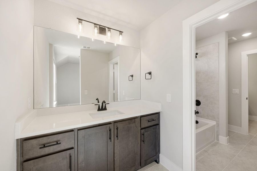 Bathroom with dark wood vanity, white countertop, large mirror, and tiled soaking tub area visible through doorway Bathroom with dark wood vanity, white countertop, large mirror, and tiled soaking tub area visible through doorway