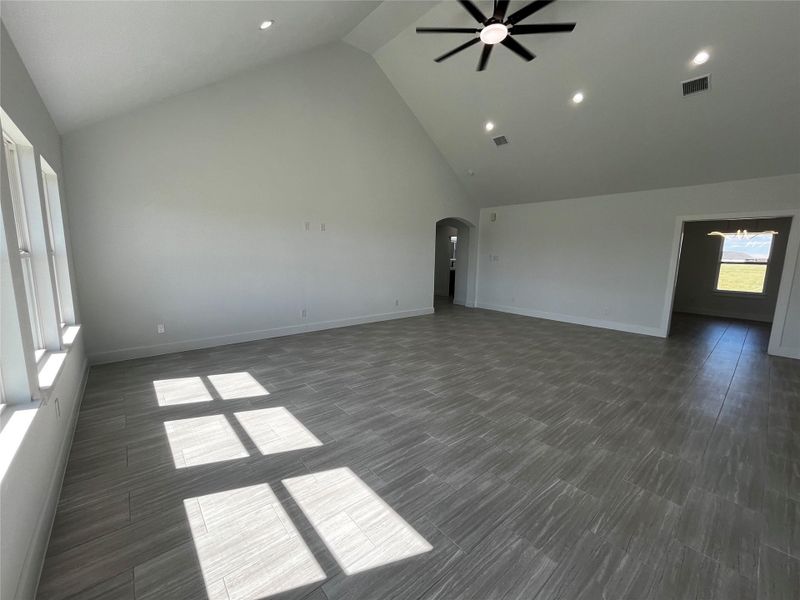 Spacious living area with high vaulted ceilings and large windows allowing ample natural light. Sleek tile flooring and modern ceiling fan enhance the contemporary feel. Adjacent room visible through an archway, offering potential for versatile use.