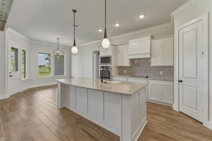 Kitchen featuring a central island with a light-toned countertop, white cabinetry, integrated stainless steel appliances, a patterned tile backsplash, and wood-finish flooring Kitchen featuring a central island with a light-toned countertop, white cabinetry, integrated stainless steel appliances, a patterned tile backsplash, and wood-finish flooring
