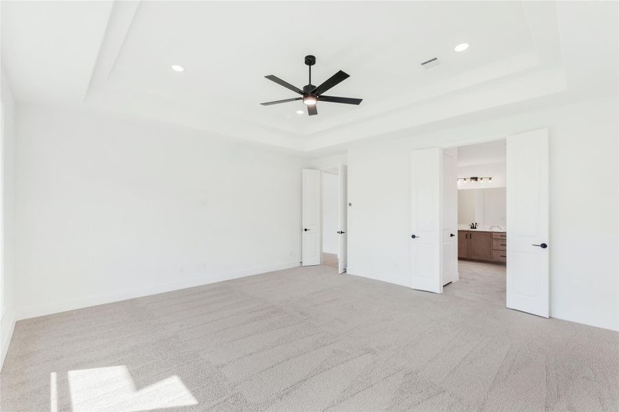 Empty primary bedroom with tray ceiling, ceiling fan, carpet flooring, and open door to ensuite bathroom